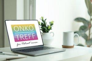 Laptop auf einem wei&szlig;en Schreibtisch zeigt ein farbiges Logo mit der Aufschrift &sbquo;ONKOTREFF f&uuml;r Patient*innen&lsquo;. Daneben stehen eine wei&szlig;e Tasse und eine kleine Pflanze.