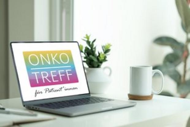 Laptop auf einem weißen Schreibtisch zeigt ein farbiges Logo mit der Aufschrift ‚ONKOTREFF für Patient*innen‘. Daneben stehen eine weiße Tasse und eine kleine Pflanze.