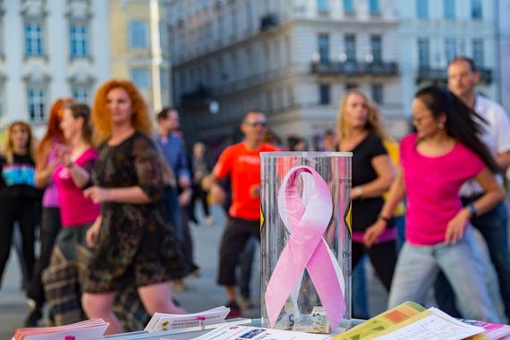 Im Vordergrund sieht man eine Spendenbox mit dem Pink Ribbon darauf, im Hintergrund tanzende Menschen. 