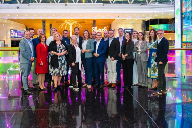 Drei Personen stehen auf der Bühne in einem Kinosaal, während eine Präsentation im Hintergrund läuft. Der Kinosaal ist mit auffälligem, pinkfarbenem Teppichboden und gelben Stühlen ausgestattet. Die Person in der Mitte hält ein Mikrofon und spricht, während die anderen beiden Personen neben ihr stehen. Auf der Leinwand ist das Logo eines Projekts zu sehen. Es ist das Team des RC Pöstlingbergs.