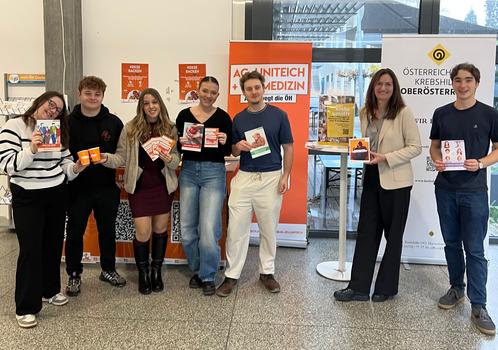 Eine Gruppe von sieben Personen steht in einem Innenraum und hält verschiedene Informationsbroschüren und Flyer in der Hand. Sie stehen vor einem Stand mit einem orangefarbenen Banner mit der Aufschrift „AG-UNITECH + AG MEDIZIN“.