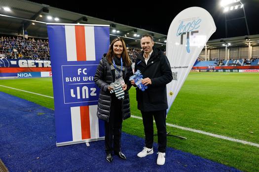 Auf dem Bild stehen Maria Sauer und ein Mann nebeneinander, beide halten kleine, gestrickte Puppen in Blau und Weiß, die Kummerkuschler. Im Hintergrund ist das Banner des FC Blau-Weiß Linz sowie das "loose TIE"-Logo zu sehen, das auf eine Charity-Aktion zugunsten der österreichischen Krebshilfe hinweist.