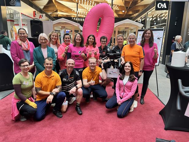 Gruppenfoto von Teilnehmenden und Unterstützer*innen einer Charity-Cycling-Aktion für Brustkrebsbewusstsein. Viele tragen pinke Kleidung, im Hintergrund steht eine große pinke Schleife.
