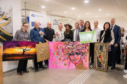 Gruppenfoto von den Gästen der Vernissage, die gemeinsam mit den Künstlern und Veranstaltern vor ihren Kunstwerken posieren. Im Vordergrund wird ein Spendenscheck in Höhe von 2.100 Euro präsentiert, umrahmt von verschiedenen Kunstwerken, die Teil der Ausstellung sind.