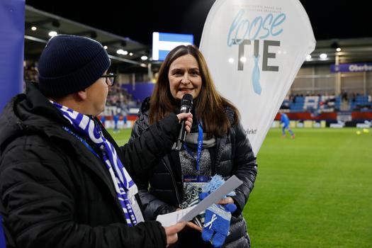 Während eines Fußballspiels von FC Blau-Weiß Linz wird Maria Sauer interviewt, die einen Mikrofonhalter in der Hand hält. Sie trägt eine schwarze Jacke und einen Schal und wird von einem männlichen Moderator unterstützt, der ebenfalls einen Wintermantel trägt. Im Hintergrund sind die Fußballspieler auf dem Spielfeld zu sehen. Das Interview bezieht sich auf eine Aktion zugunsten der österreichischen Krebshilfe, was durch das Banner mit der Aufschrift "loose TIE" verdeutlicht wird. Die Frau hält eine kleine, handgefertigte, blaue Puppe, die als Teil der Charity-Aktion präsentiert wird.