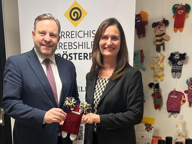 Mag. Franz Ebner und Gesch&auml;ftsf&uuml;hrerin Maria Sauer halten gemeinsam einen Kummerkuschler und posieren vor dem Plakat der Krebshilfe O&Ouml;