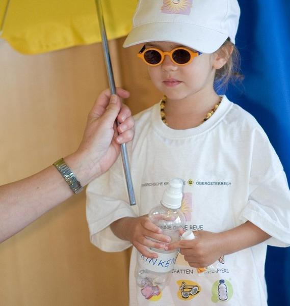 M&auml;dchen im Kindergartenalter, das eine Kappe, eine Sonnenbrille und ein zu gro&szlig;es wei&szlig;es T-Shirt tr&auml;gt mit den Sonnenregeln darauf. In der Hand h&auml;lt sie eine leere Plastikwasserflasche und eine kleine Sonnencremeprobe. &Uuml;ber ihr wird ein kleiner Sonnenschirm gehalten. 