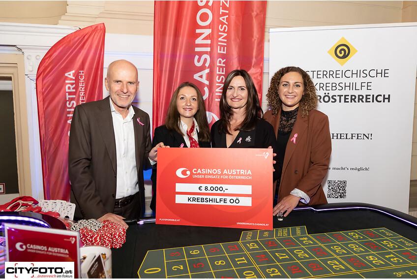 Das Bild zeigt eine feierliche Übergabe eines Schecks über 8.000 Euro von Casinos Austria an die Krebshilfe Oberösterreich. Vier Personen stehen zusammen an einem Roulettetisch: Ein Mann in Anzug und rosa Schleife, zwei Frauen in eleganter Kleidung und eine weitere Frau mit einer Krawatte, die sich für den guten Zweck engagieren. Der Hintergrund zeigt Banner von Casinos Austria und der Österreichischen Krebshilfe. Alle tragen zur Unterstützung der Krebsbekämpfung bei.