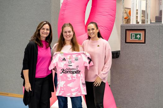 Drei Frauen in pinker Kleidung halten gemeinsam das pinkfarbene Trikot der LINZ AG Steelvolleys, das im Rahmen eines Events zur Brustkrebsaufklärung präsentiert wird. Der Hintergrund zeigt einen rosa Luftbogen.