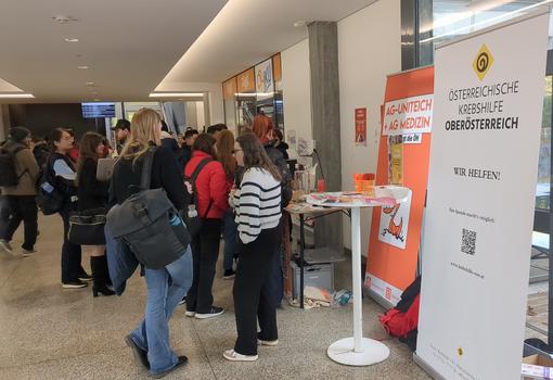 Eine größere Gruppe von Menschen steht in einem Innenraum und sieht aus, als ob sie an einem Informationsstand interessiert sind. Der Stand hat ein großes Banner mit der Aufschrift „ÖSTERREICHISCHE KREBSHILFE OBERÖSTERREICH“ und der Slogan „WIR HELFEN!“